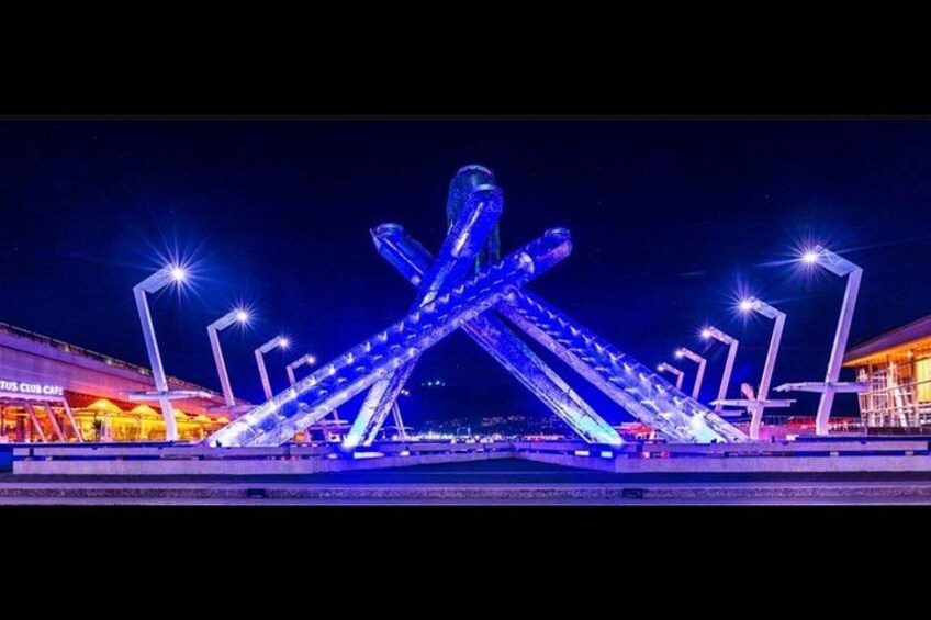 A brightly lit sculpture with intersecting arms, a shopping area in the background, and a clear night sky. 2