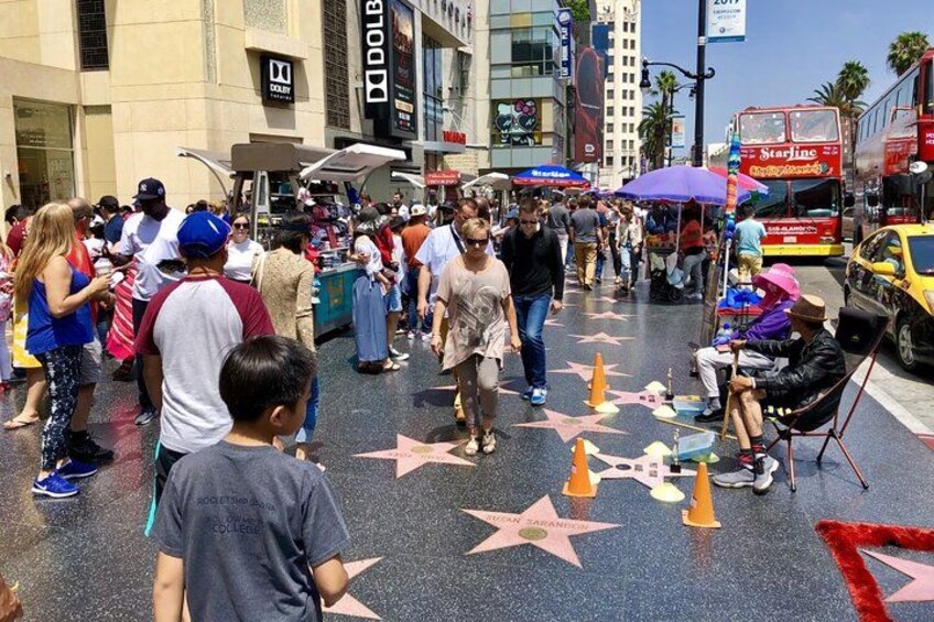 Une rue animée où l’on voit des gens qui marchent et qui sont à l’arrêt, un autobus Starline et le « Hollywood Walk of Fame ». 1