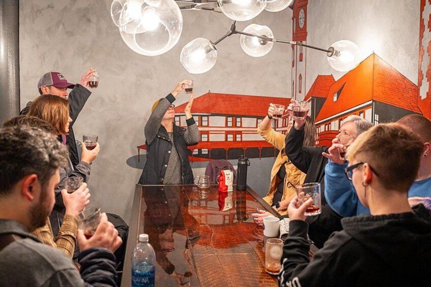 Een groep mensen proost met drankjes rond een tafel in een ruimte met een muurschildering van een gebouw. 2