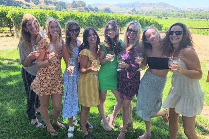 A group of people standing in a vineyard with rows of grapevines in the background. 5