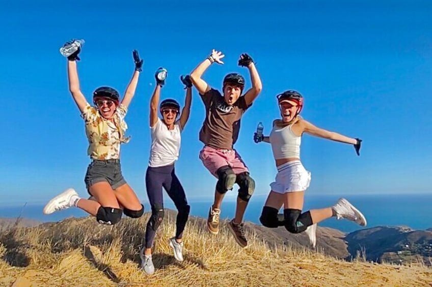 Four people jumping on a grassy hill with a clear sky and distant mountains. 5