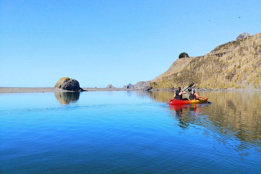 Dos personas haciendo kayak en aguas tranquilas, con una roca grande al frente y un paisaje montañoso al fondo. 3