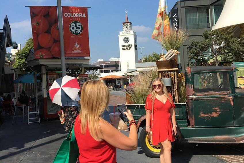 Une femme vêtue d’une robe rouge se tient à côté d’un camion ancien sur lequel est posée une plante. On aperçoit également un étal de marché avec un parasol coloré et une grande banderole rouge portant un texte. 3