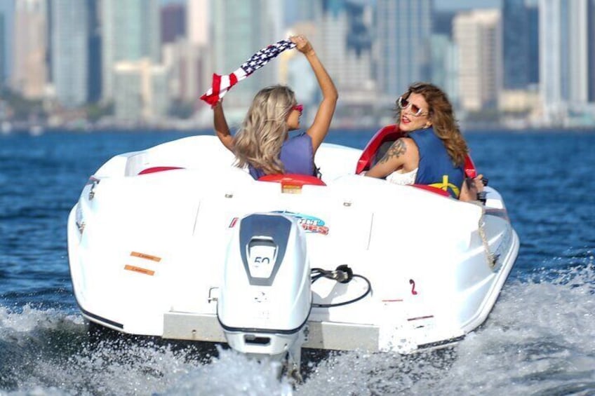 Un jet ski bianco con due persone a bordo, una delle quali tiene una bandiera americana, che si muove sull'acqua con uno skyline cittadino sullo sfondo. 1