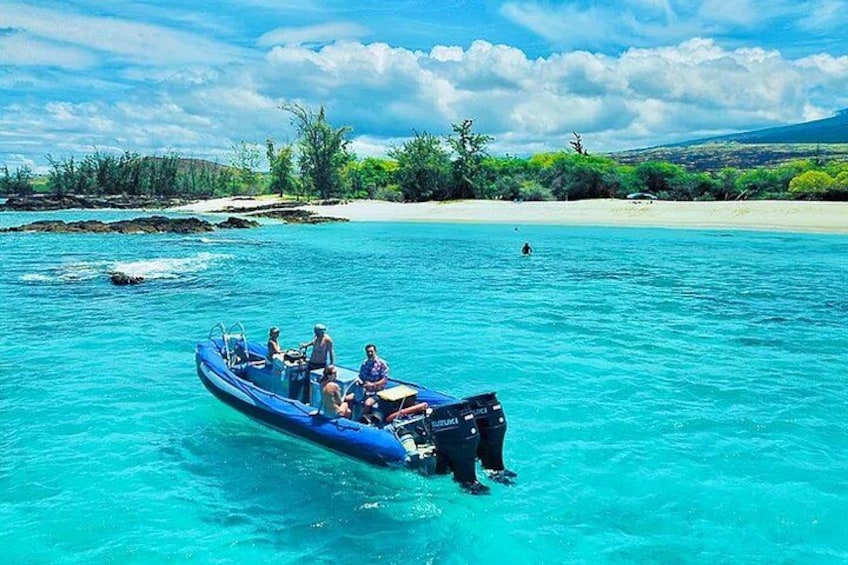 Ein blaues Boot mit Personen auf einem türkisblauen Meer, ein Sandstrand und grünes Ufervegetation im Hintergrund. 3