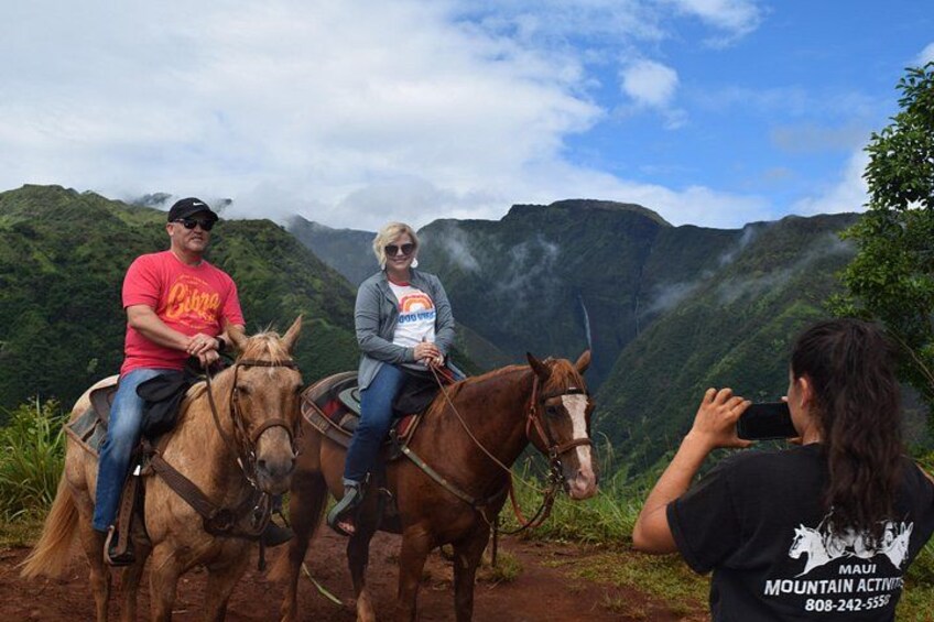Two people riding horses on a mountain trail with lush greenery and a clear sky. 1