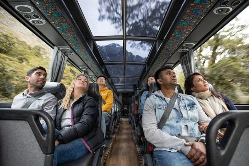 Ein Businnenraum mit sitzenden Fahrgästen, einer Karte an der Decke und Blick auf Bäume und Berge vor den Fenstern. 2