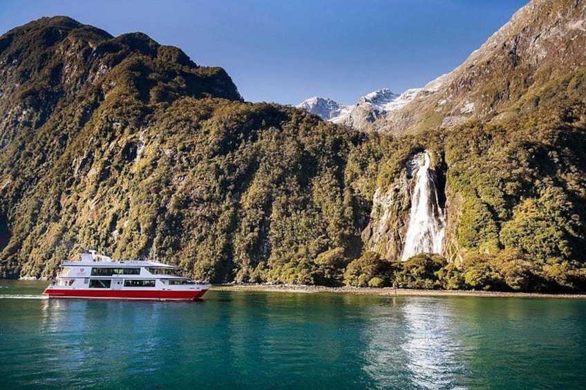 Ein Boot auf einem ruhigen See mit einem Wasserfall und bewaldeten Bergen im Hintergrund. 1