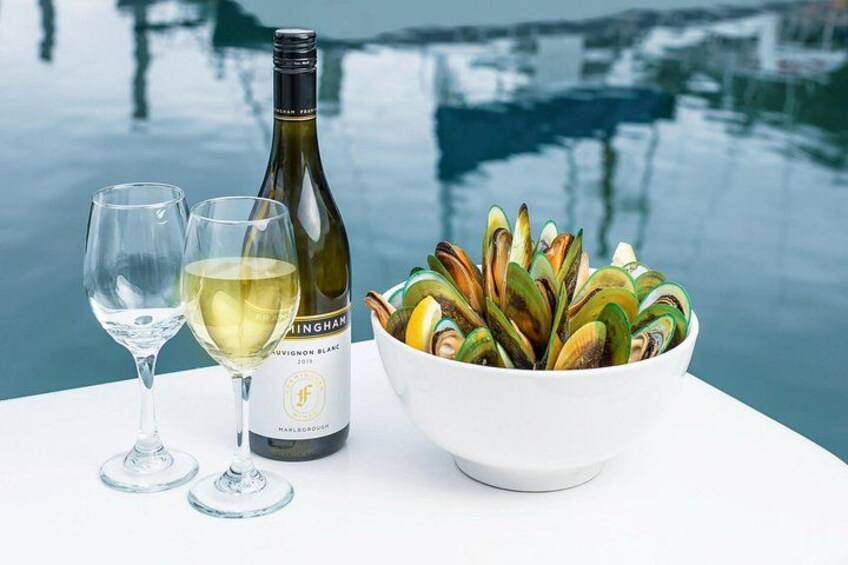 Una botella de vino, dos copas y un bol de mejillones sobre una mesa junto al agua. 3
