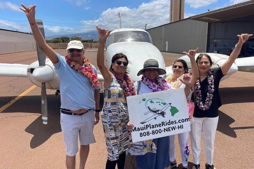 A group of people standing in front of an airplane with a sign that reads 'MauiPlaneRides.com 808-800-NEW-HI'. 2