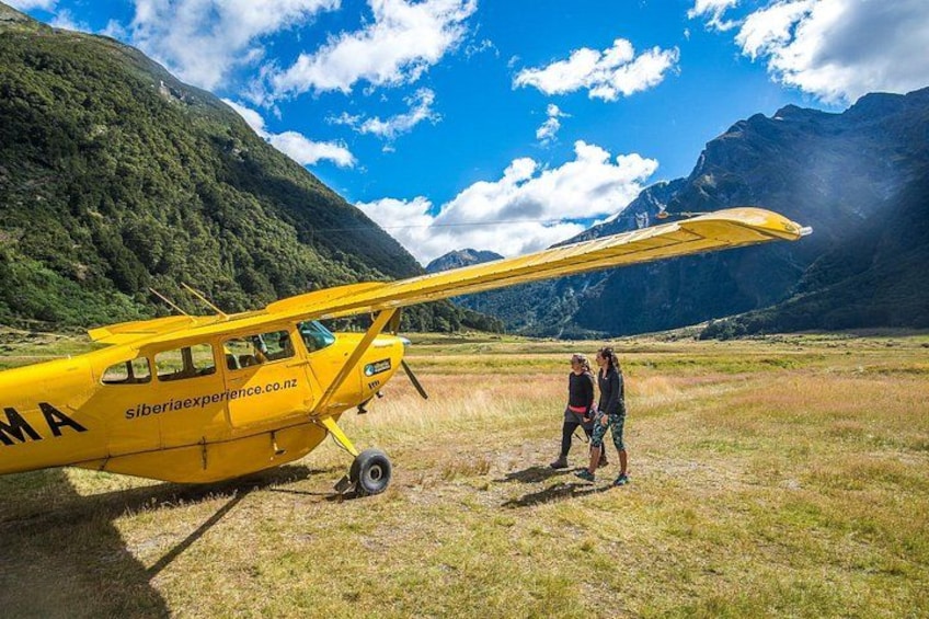 A yellow airplane with 'Siberia Experience' written on it, parked on a grassy field with mountains in the background. 1