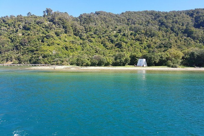 A lake with clear blue water, a small white structure on the shore, and a forested hill in the background. 1