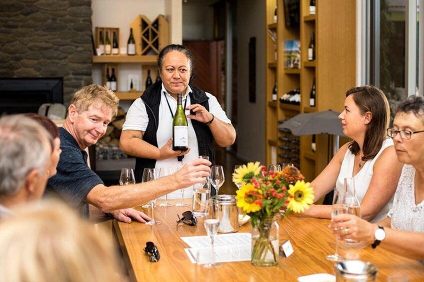 Un grupo de personas sentadas alrededor de una mesa de madera con copas de vino, una botella de vino y un jarrón con girasoles. 1