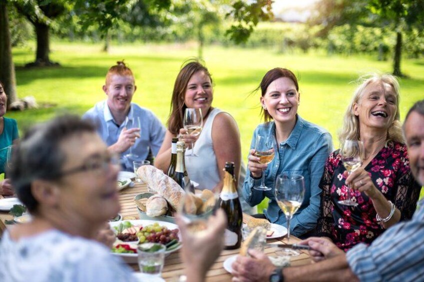 Un groupe de personnes est assis à une table à l’extérieur, devant de la nourriture et des boissons, dans un espace verdoyant. 1