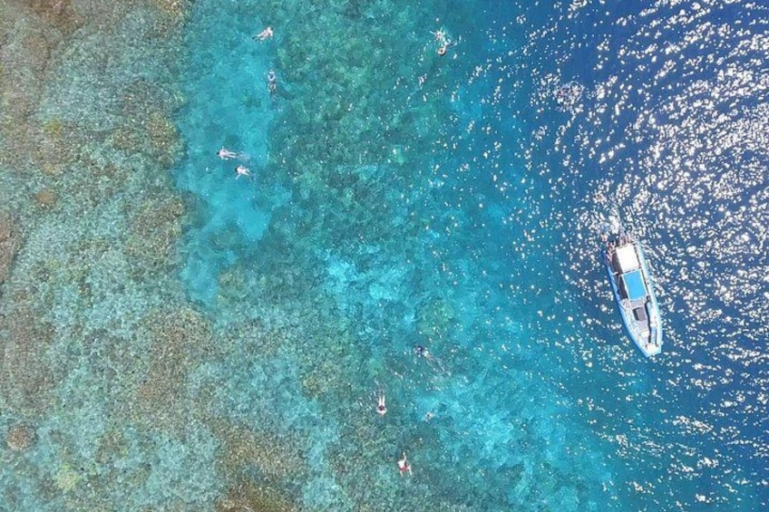 A boat with three people swimming in clear blue water, surrounded by a coral reef. 4