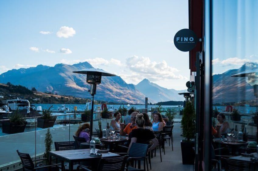 Ein Restaurant am Seeufer mit Außenbestuhlung, Bergen im Hintergrund und blauem Himmel. 2
