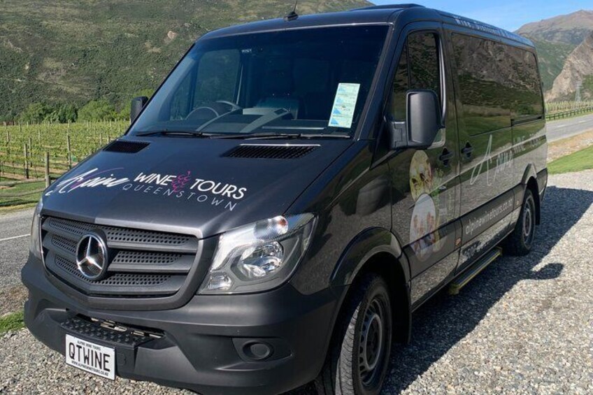 Ein schwarzer Van mit der Aufschrift 'WINE TOURS QUEENSTOWN' steht auf einem Schotterweg vor einer Bergkulisse. 3