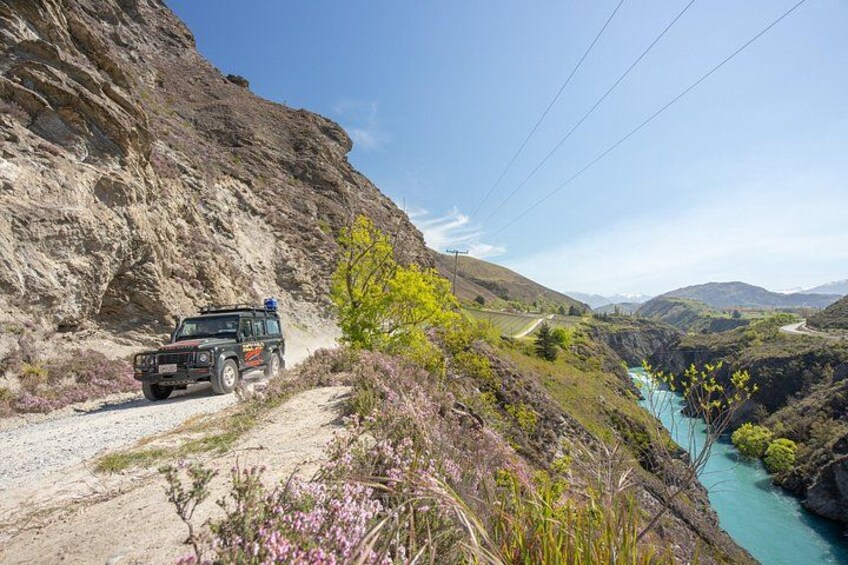 A vehicle on a gravel path beside a river, with a rocky cliff and greenery in the background. 2