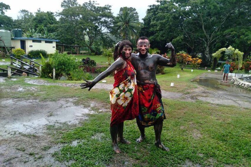Dos personas con vestimenta tradicional de pie en un área cubierta de césped, con un estanque y árboles al fondo. 5