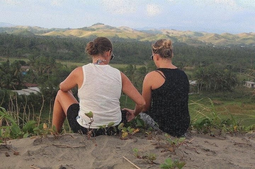 Two people sitting on a sandy hill overlooking a hilly landscape. 1