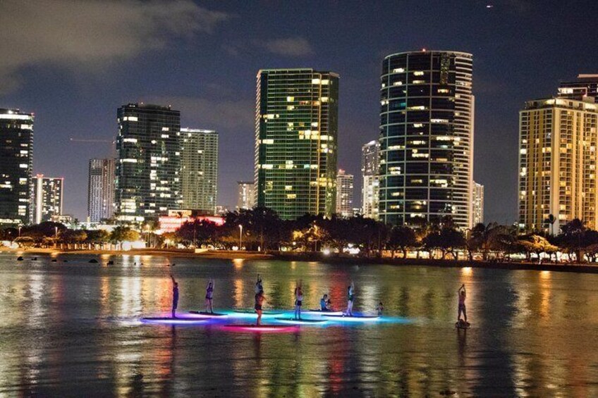 Panorama notturno di una città con edifici illuminati e un gruppo di persone su una piattaforma galleggiante in acqua. 3