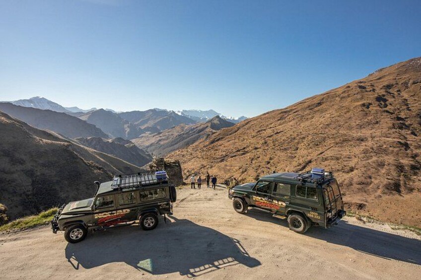 Two off-road vehicles parked on a dirt road in a mountainous area with a clear sky. 1