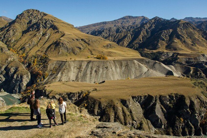 A group of people standing on a rocky outcrop overlooking a vast, rugged landscape with layered hills and a clear sky. 5