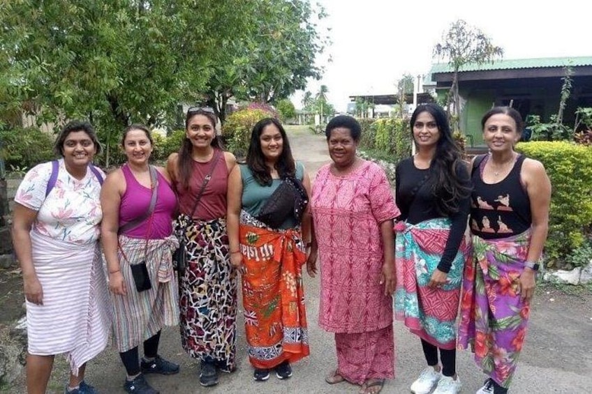 Un grupo de personas de pie al aire libre, vestidas con atuendos tradicionales y casuales de colores variados, con follaje y un edificio al fondo. 5