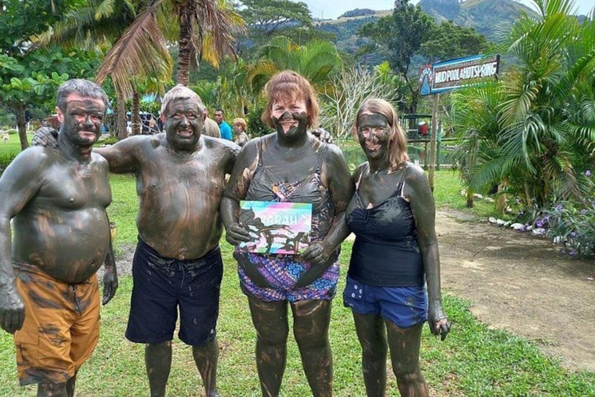 Cuatro personas cubiertas de barro, de pie en un parque con un letrero que dice 'Mud Pit Challenge'. 2