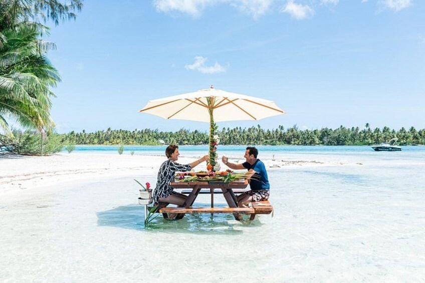 Dos personas sentadas en una mesa de madera en una playa con una sombrilla grande, agua cristalina y palmeras al fondo. 4