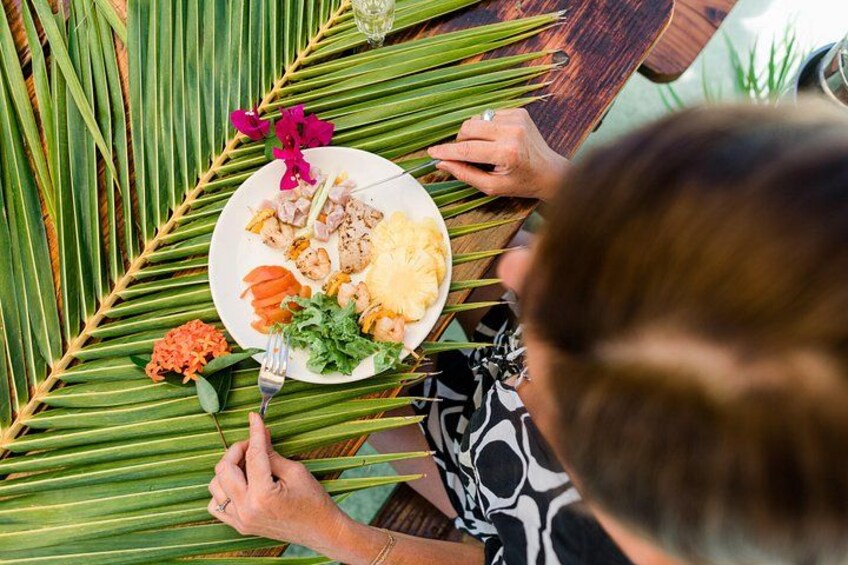 Una persona sentada a una mesa con un plato de comida, hojas de palma y una bebida. 2