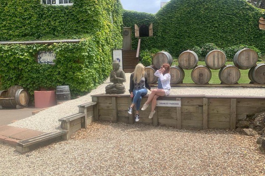 Twee personen zitten op een houten bank, met een stenen beeld en houten vaten op de achtergrond. 5