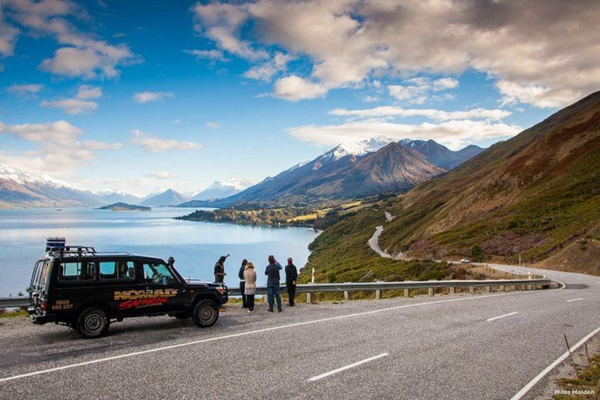 Ein schwarzes Fahrzeug mit Aufschrift 'NORAD' steht an einer Straße, daneben Personen, eine kurvenreiche Straße, ein See und Berge im Hintergrund. 1