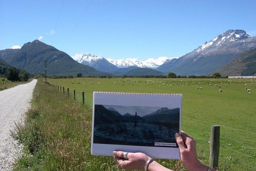 A person holding a tablet displaying a mountain landscape, a gravel road, and a green field with sheep. 3