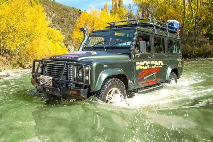 Ein Land Rover fährt durch einen Fluss, umgeben von Herbstbäumen. 4