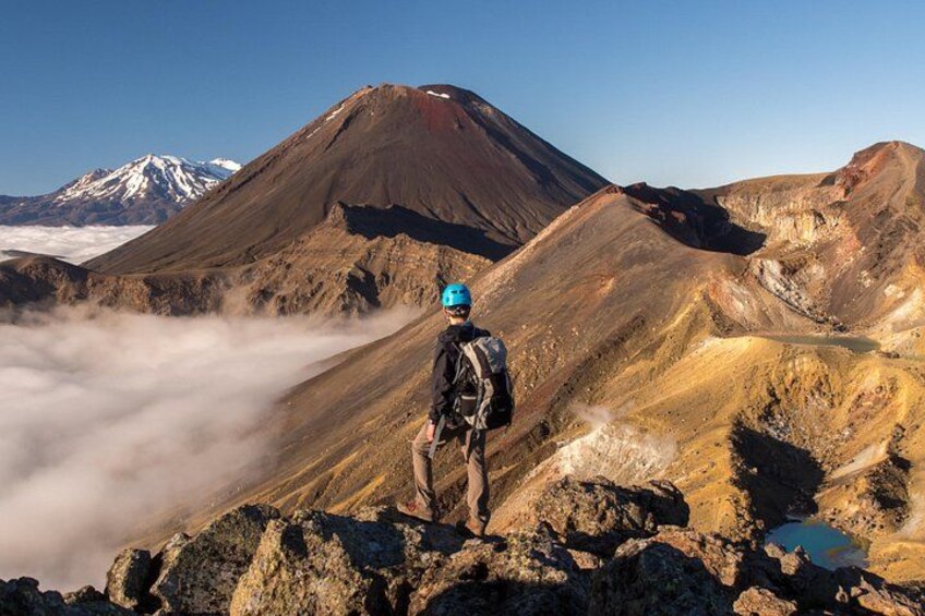 A hiker on a rocky mountain peak with a view of a volcano and clouds below. 3