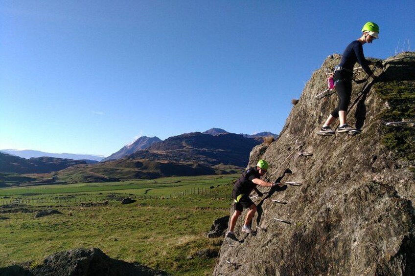 Two people climbing a rocky hill with a scenic landscape in the background. 5