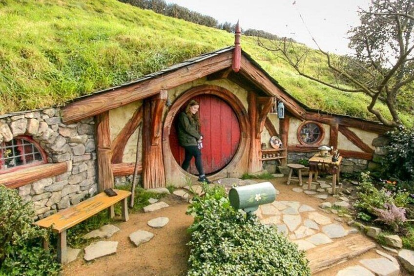 Una casa de estilo Hobbit con puerta redonda, paredes de piedra y una colina cubierta de hierba al fondo. 1