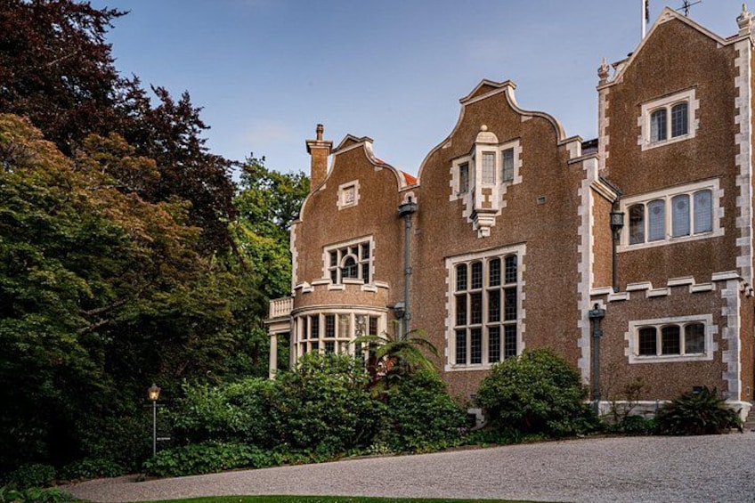 A large brick building with multiple windows and a turret, surrounded by lush greenery. 1