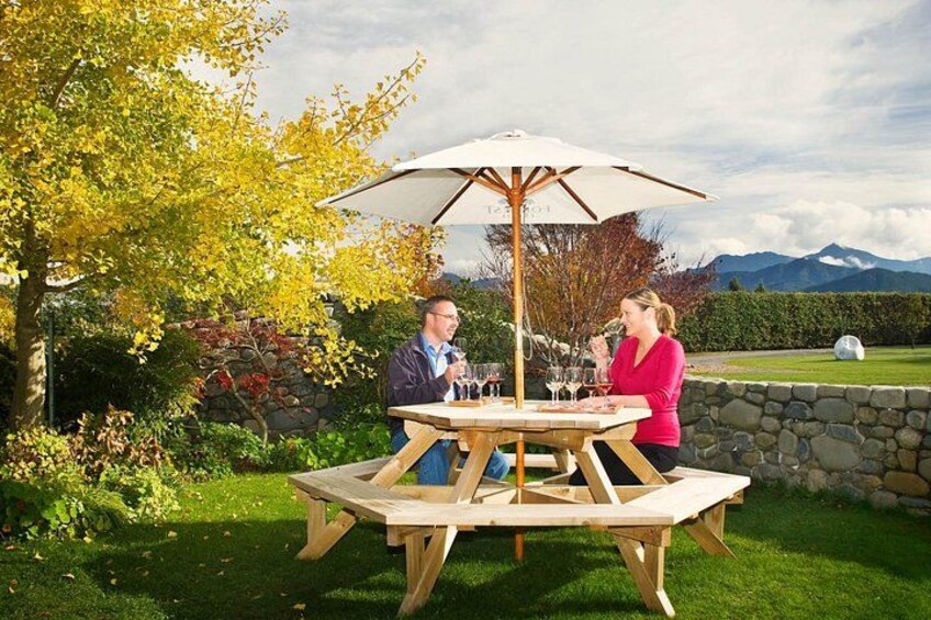 Een houten picknicktafel met een parasol, twee personen zittend, en een bergachtig landschap op de achtergrond. 5