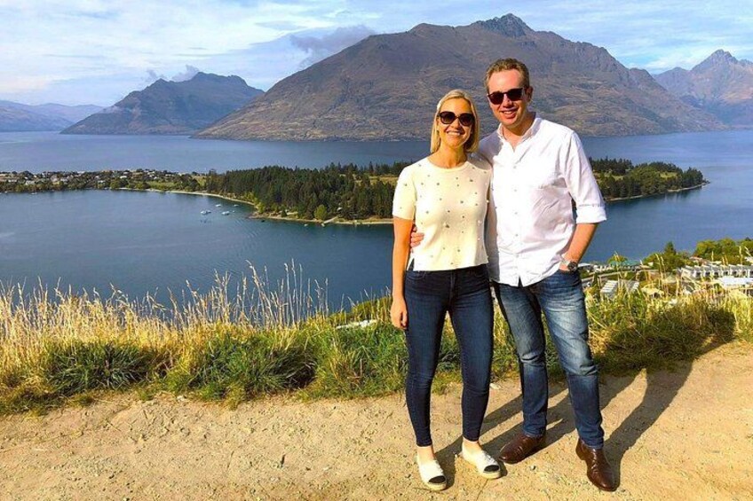 A couple standing on a dirt path with a lake and mountains in the background. 2