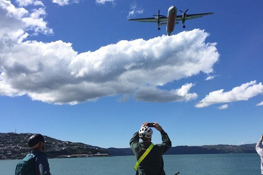 Eine Person fotografiert ein Flugzeug, das über ein Gewässer mit Bergen im Hintergrund fliegt. 2