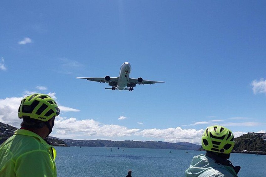 Eine Person mit Helm und einer leuchtend gelben Jacke beobachtet ein Flugzeug, das über dem Wasser fliegt. 3