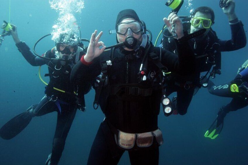 Four scuba divers underwater, wearing black wetsuits and scuba gear, with one diver making a peace sign. 5