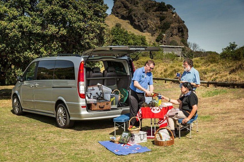 Un minivan argentato parcheggiato sull'erba con il bagagliaio aperto, un'attrezzatura da picnic con tovaglia rossa e una collina rocciosa sullo sfondo. 4