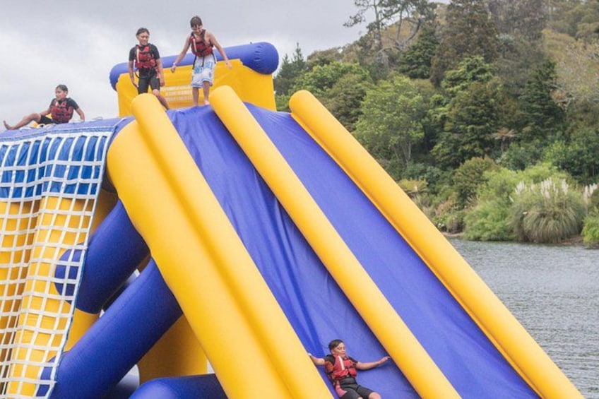 A water slide with blue and yellow colors, surrounded by greenery and a body of water. 1