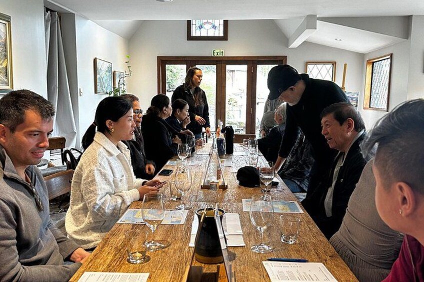 A group of people seated around a long wooden table with wine glasses and bottles, in a room with a door and windows. 4