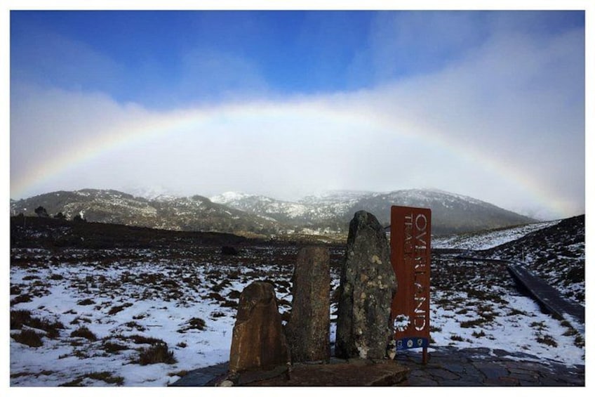 Paisaje nevado con un arcoíris, un cartel de indicaciones y montañas al fondo. 1