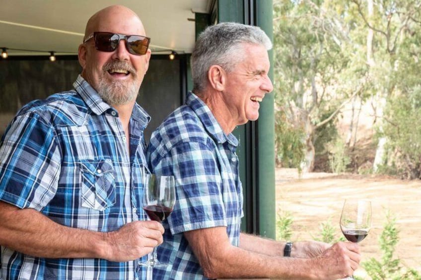 Two men in plaid shirts holding wine glasses, standing outdoors with trees in the background. 1