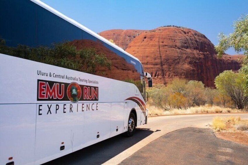 A white bus with 'EMU RUN EXPERIENCE' and 'Uluru & Central Australia Touring Specialists' written on it, parked on a road with a red rock formation in the background. 2